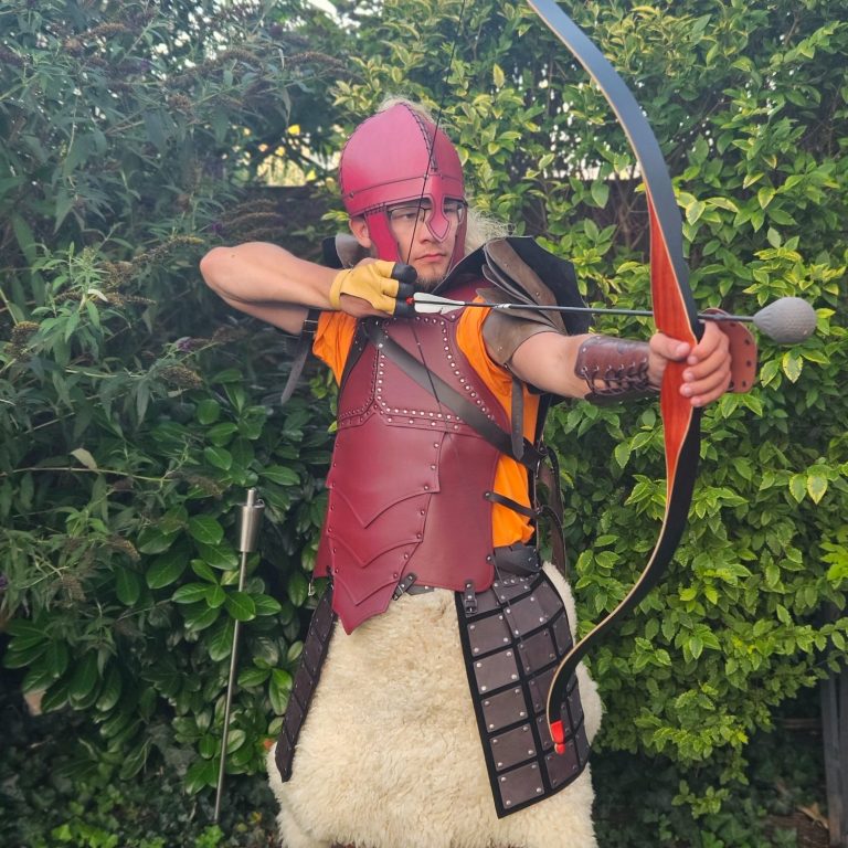 Krieger in historischer Rüstung mit Bogen, vor einer grünen Hecke.