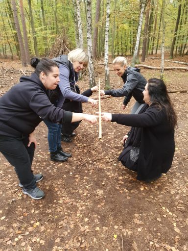 Vier Personen in einem Wald, die gemeinsam einen Stock halten und sich gegenüberstehen.