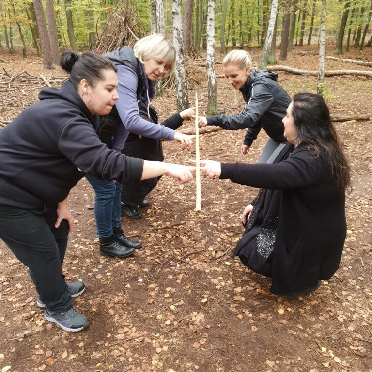 Vier Frauen im Wald arbeiten zusammen, um einen langen Stock balanciert zu halten.