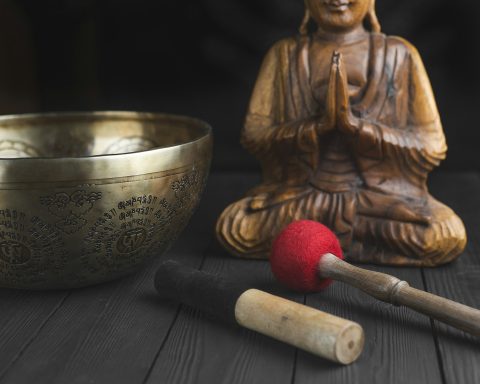 Holzkörper eines Buddhas neben einer Klangschale und Schlägern auf dunklem Untergrund.