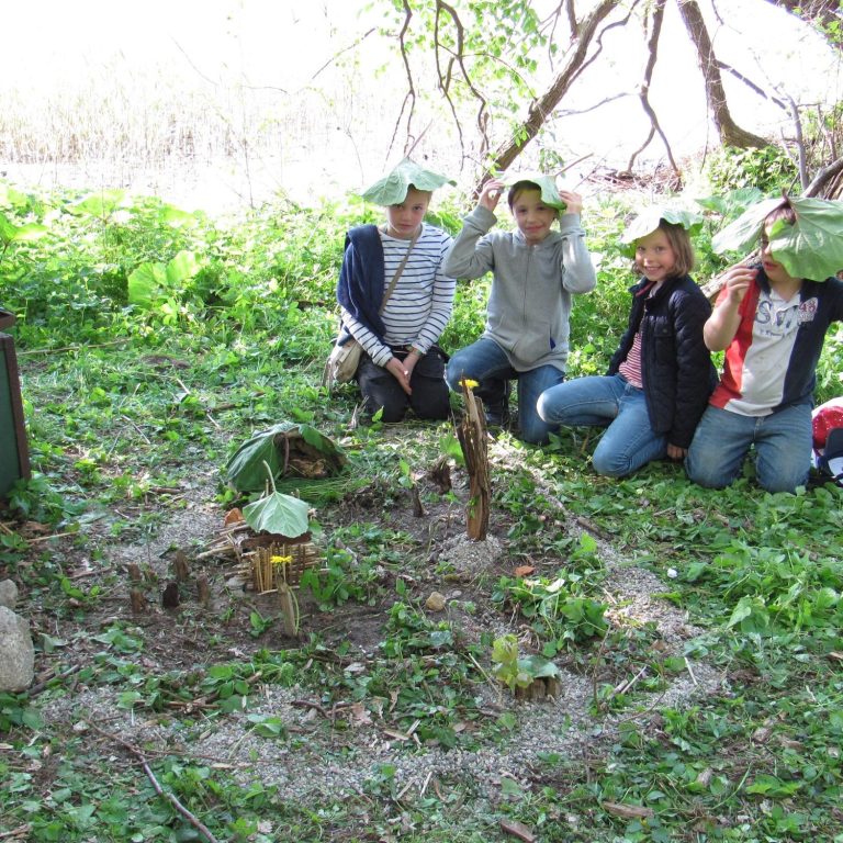 Gruppe von fünf Personen mit Blättern auf dem Kopf, die in einer Waldlichtung sitzen.