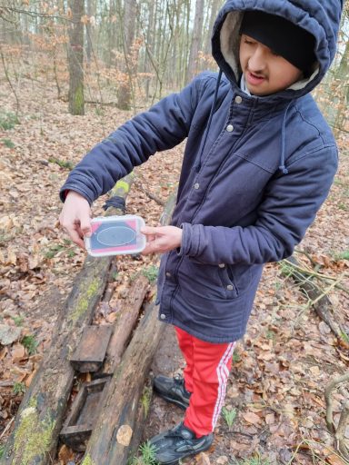 Junger Mann im Wald hält eine Box in den Händen, trägt eine Jacke und rote Hose.