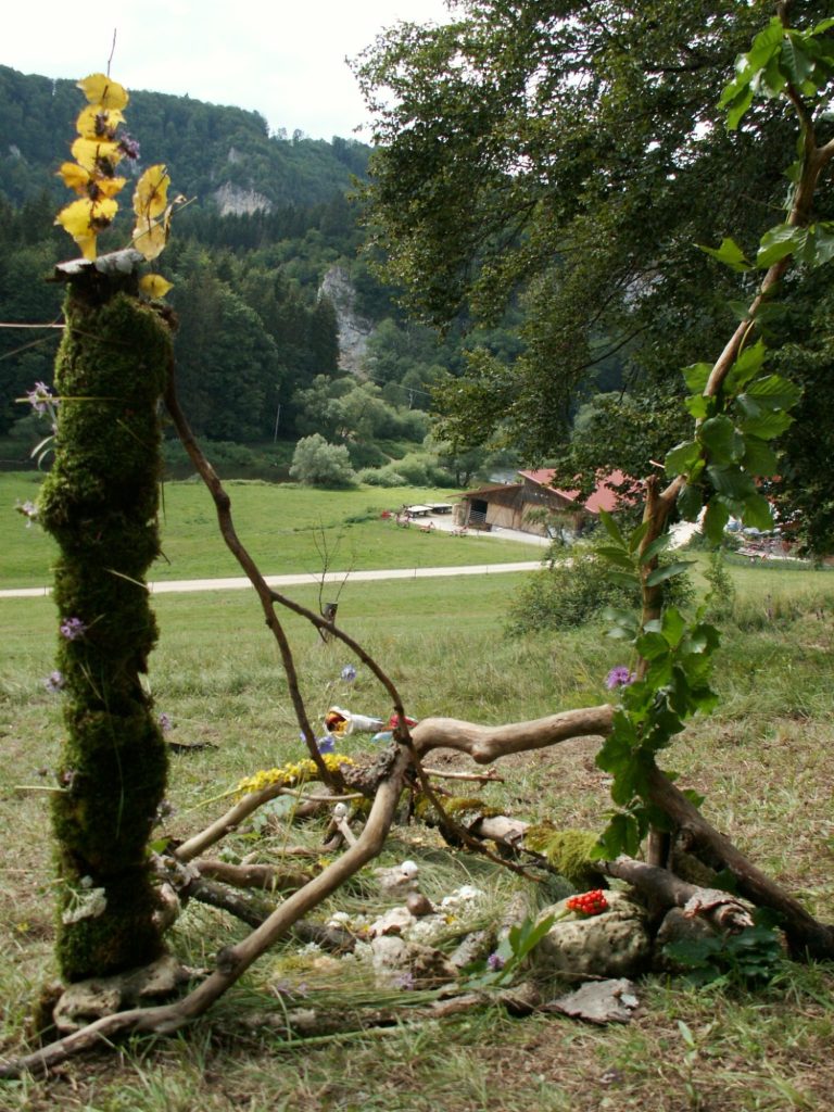 Naturinstallation mit moosbedeckten Stämmen, Blumen und Zweigen in einer grünen Landschaft.