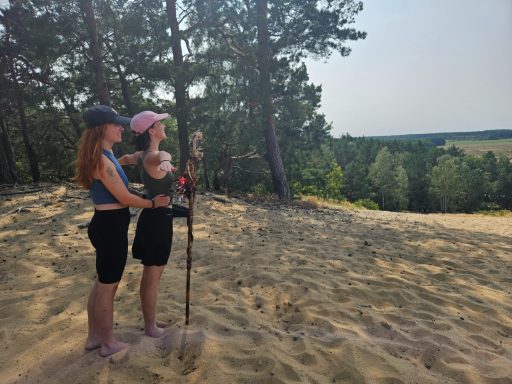 Zwei Frauen stehen auf einem Sandhügel, umgeben von Bäumen und Landschaft.