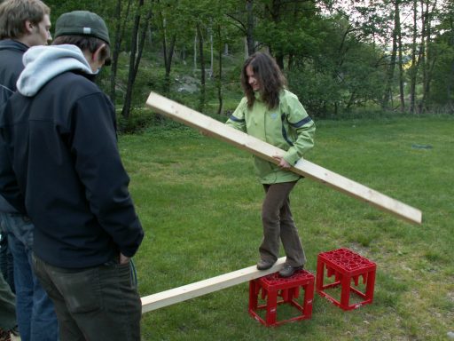 Eine Person balanciert auf einer Holzplanke zwischen zwei roten Kästen im Freien.