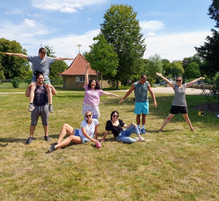 Sechs Personen posieren im Freien, auf einer Wiese unter blauem Himmel.