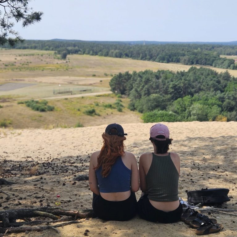 Zwei Personen sitzen auf einem Hügel und blicken auf eine weite Landschaft.