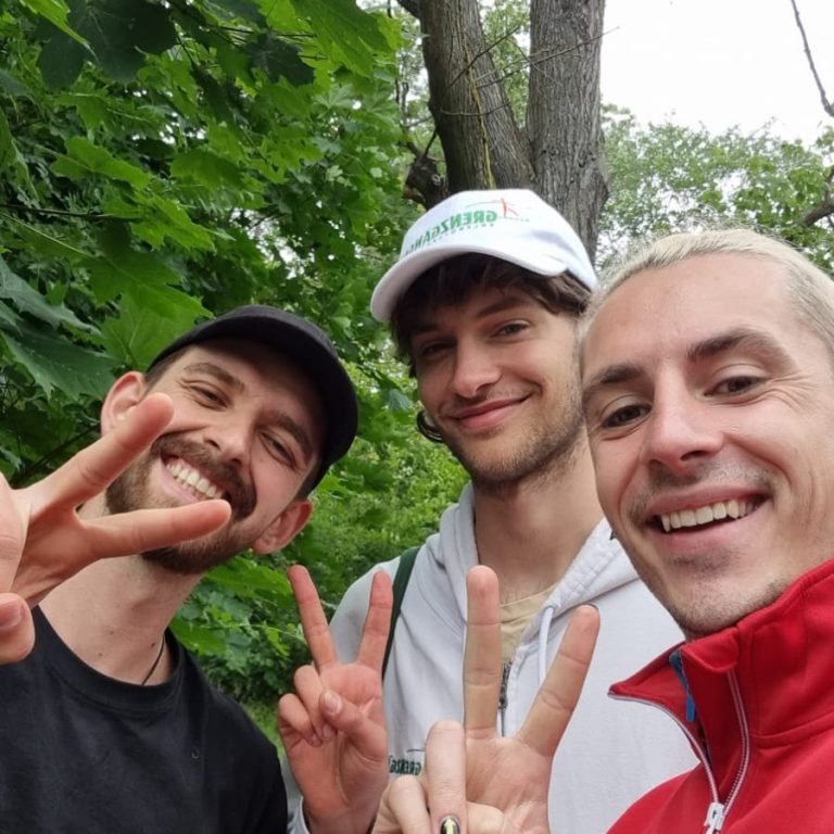 Drei Männer posieren im Freien und zeigen das Peace-Zeichen. Hintergrund mit grünen Blättern.