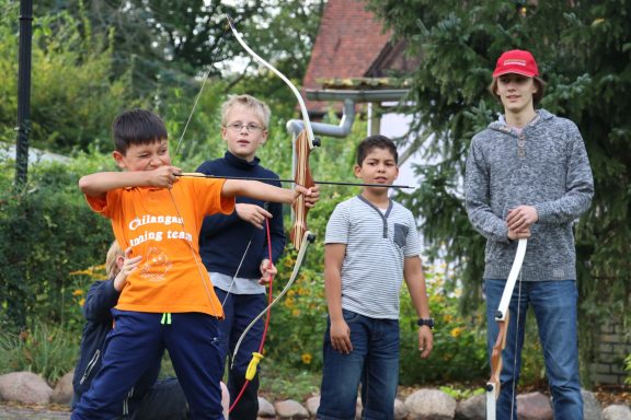 Vier Jungen stehen im Freien, während einer mit einem Bogen zielt. Blumen im Hintergrund.