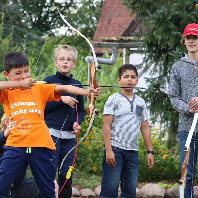 Fünf Kinder stehen im Freien, zwei halten Bögen, während andere zuschauen.