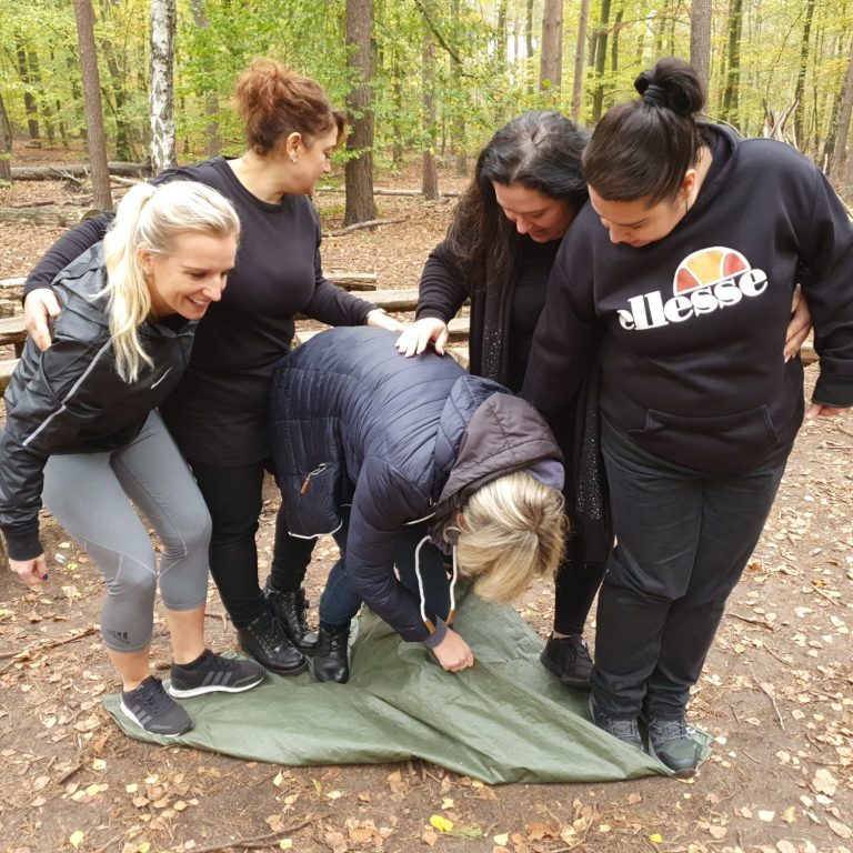 Fünf Frauen stehen in einem Wald, eine kniet auf einer Decke und die anderen helfen.