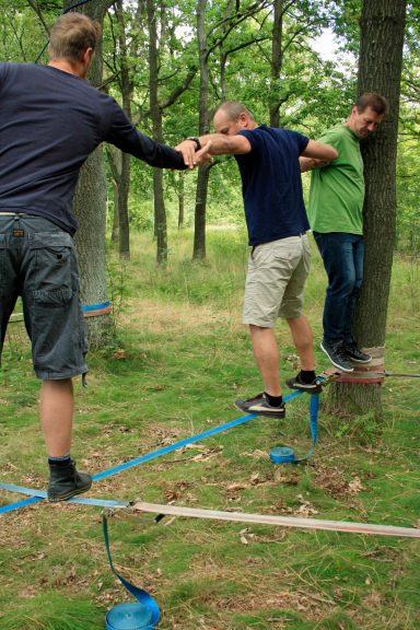 Drei Personen balancieren auf einem Slackline zwischen Bäumen in einem waldigen Gebiet.