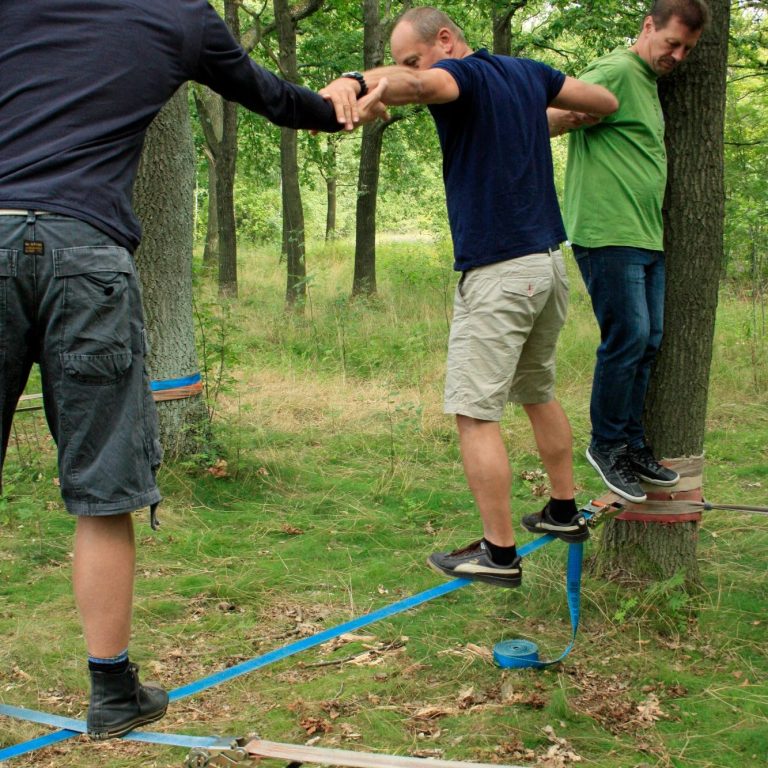 Drei Personen balancieren auf einem Slackline zwischen Bäumen im Wald.