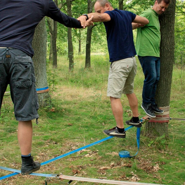 Drei Personen balancieren auf einem Slackline zwischen Bäumen im Wald.