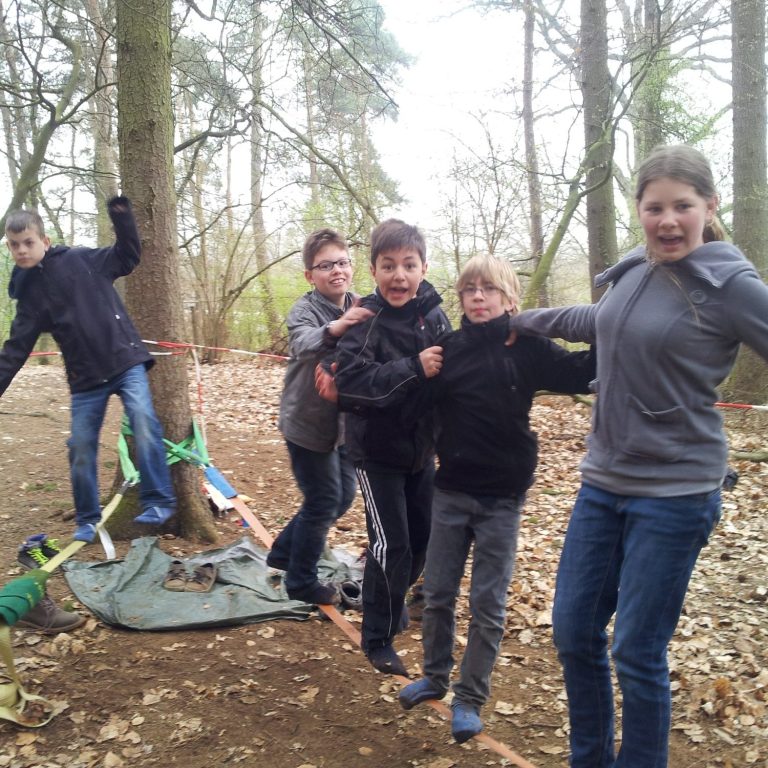 Fünf Kinder stehen in einem Wald, einige balancieren auf einem Seil zwischen Bäumen.