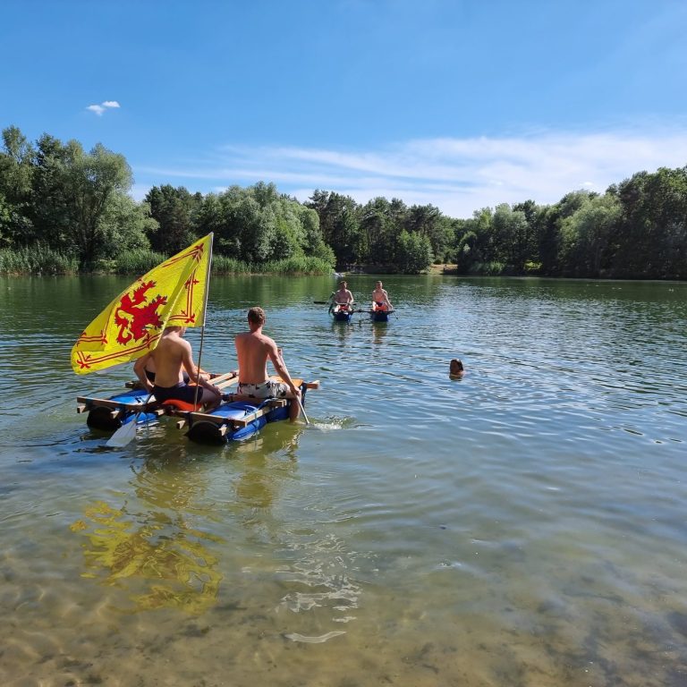 Junge Menschen paddeln auf einem Gewässer, umgeben von Bäumen und blauem Himmel.